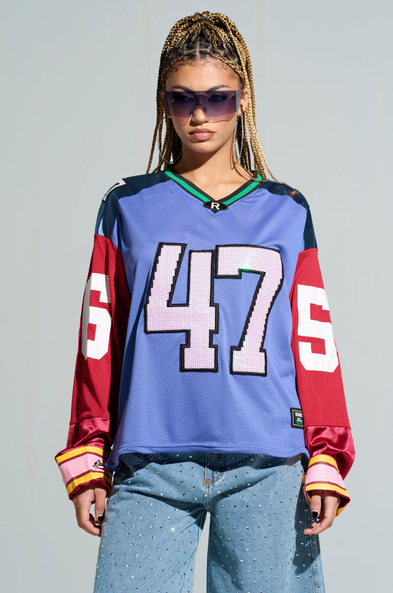 Dressed in a GAME TIME JERSEY featuring the number 47, an individual displays an oversized silhouette. The striking red sleeves with the number 55 form a bold color block pattern. With braided hair and sunglasses, they exude confidence against a plain backdrop, wearing blue jeans embellished with studs. 2