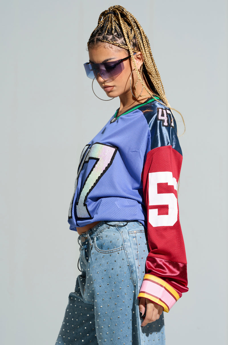 Against a neutral backdrop, someone is sporting the vibrant GAME TIME JERSEY adorned with numbers 47 and 53 on the sleeves, coupled with studded jeans. Their braided hair and oversized sunglasses perfectly capture the lively color block pattern style. 3