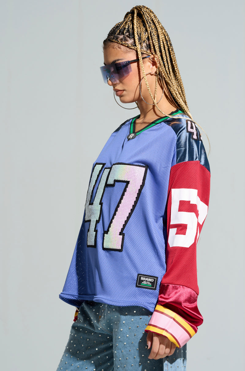 With braided hair, an individual wearing the GAME TIME JERSEY in a striking color block design of blue and red stands against a plain background. The oversized jersey displays bold numbers "47" and "57" and is paired with sunglasses and jeans embellished with small studs. 4