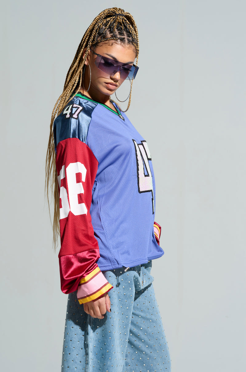 A person confidently stands against a plain background, wearing sunglasses and the GAME TIME JERSEY, known for its striking color block design. They complete their look with patterned loose blue pants, large hoop earrings, long braided hair, and an oversized silhouette. 5