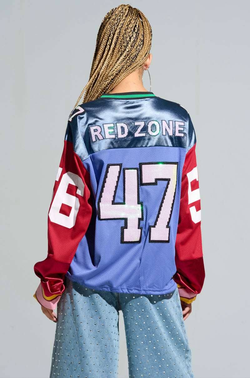 With their back to the camera, a person with braided hair showcases an oversized GAME TIME JERSEY in blue, red, and white featuring "RED ZONE" and the number 47. They pair it with light blue wide-leg jeans for a striking color block pattern look. 6