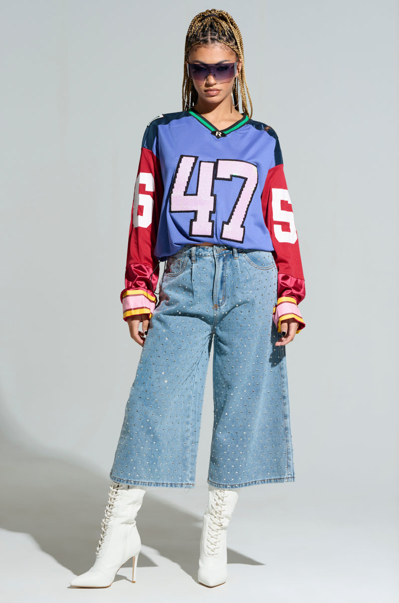 A person wearing a vibrant GAME TIME JERSEY with the number 47, paired with wide-leg denim jeans, white high-heeled boots, and sunglasses. They exude confidence in the oversized silhouette as they stand boldly against a plain background. 7