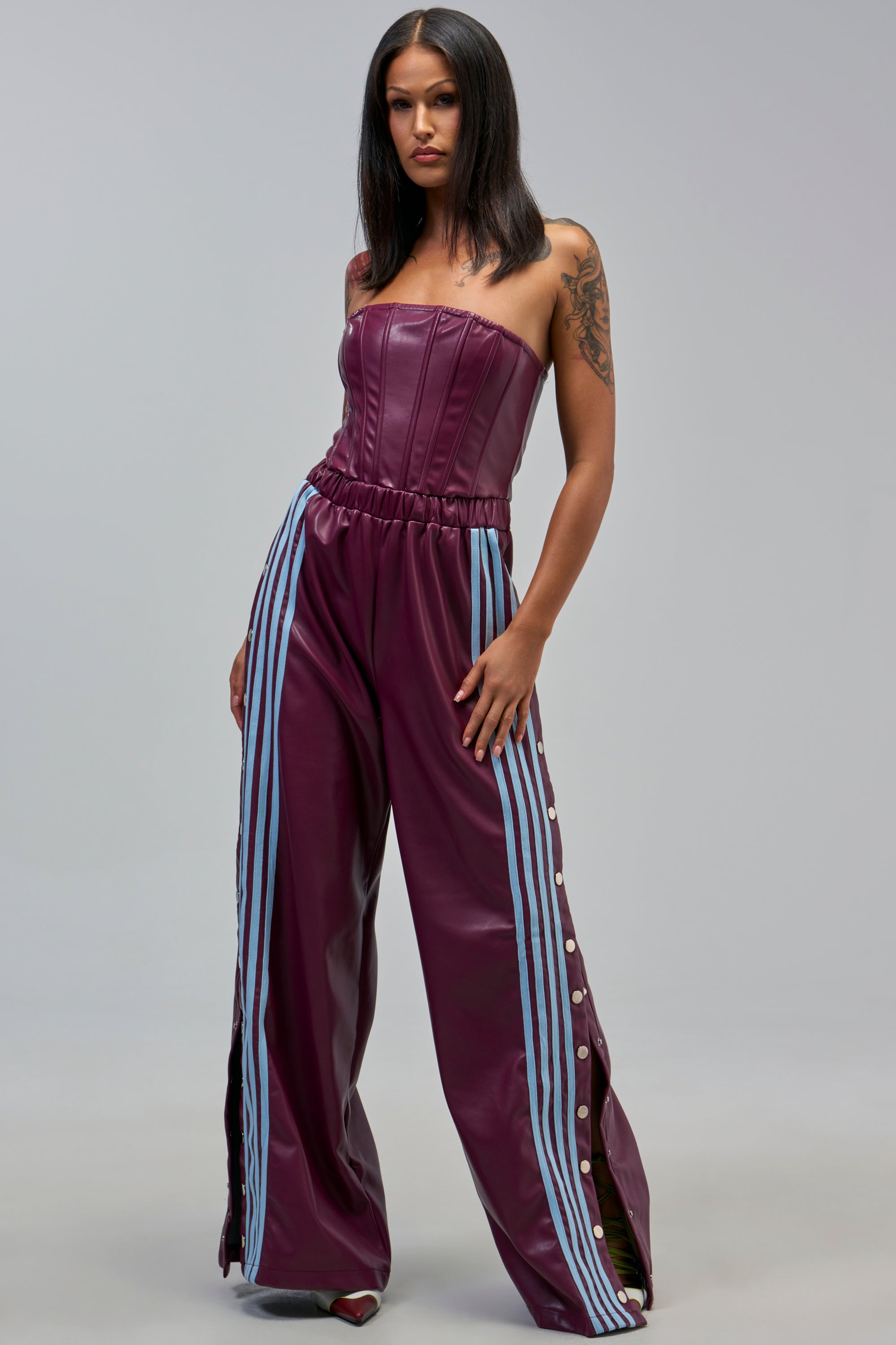 A woman stands confidently in the SEE YOU NEXT YEAR PU JUMPSUIT IN BURGUNDY, a strapless faux leather piece with light blue racing stripes and button accents, against a plain gray background.