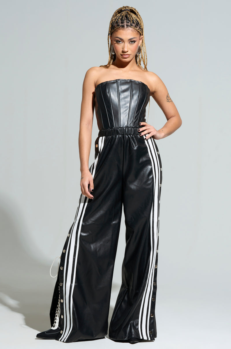 A person with braided hair is confidently posing against a plain background, wearing the SEE YOU NEXT YEAR PU JUMPSUIT, featuring wide-leg pants with white stripes and snaps on the sides.