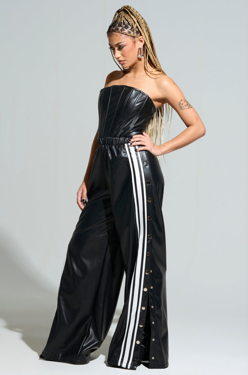A woman with braided hair exudes confidence as she stands against a plain background, looking down with one hand on her hip. She wears the chic SEE YOU NEXT YEAR PU JUMPSUIT, featuring a strapless faux leather top and wide-leg black pants with white stripes and side buttons.