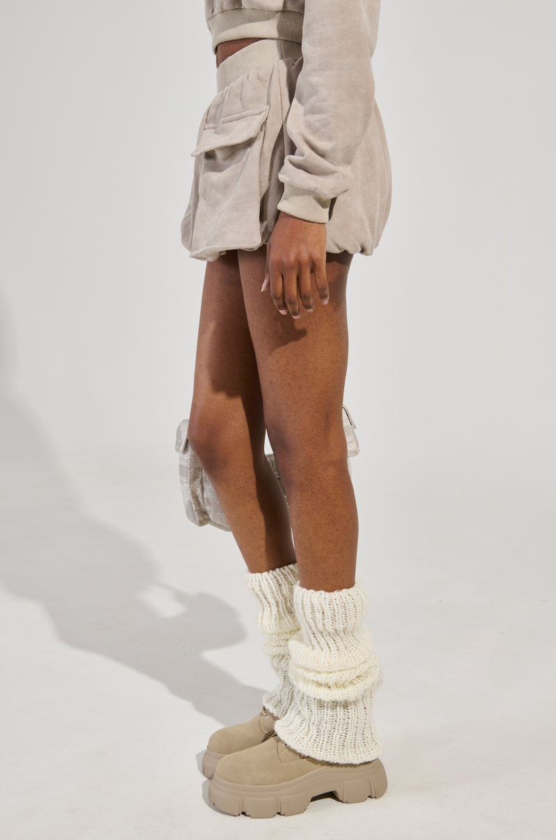 A person wearing an OFF-DUTY MINI SKIRT as part of a beige outfit, with a cropped long-sleeve top and bubble hem puffy shorts, paired with chunky beige ankle boots and white knitted leg warmers, stands against a plain background. Their right hand is slightly visible by their side.