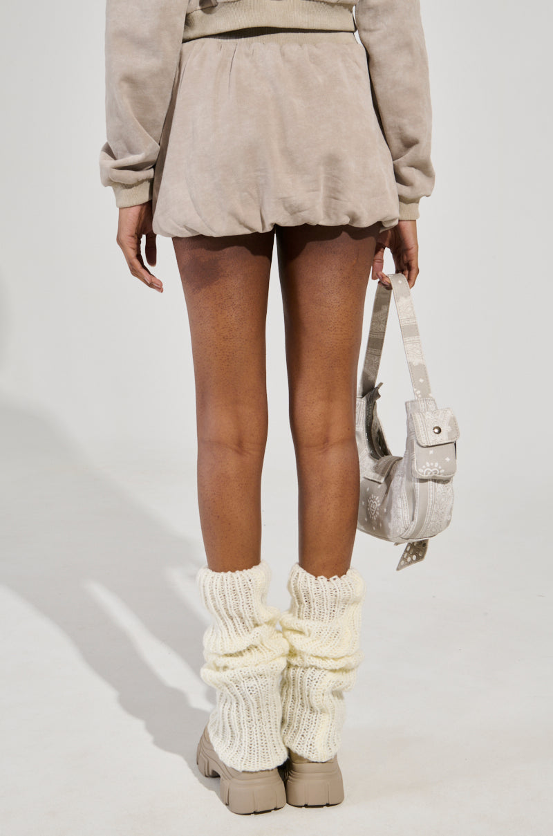 A person stands with their back to the camera, wearing the OFF-DUTY MINI SKIRT in light beige with a matching top and holding a small handbag. Cream-colored knit leg warmers accentuate the light shoes against a plain, neutral backdrop.