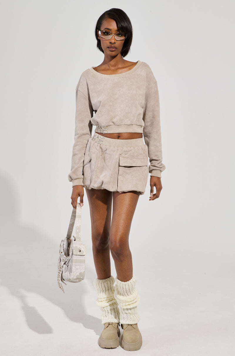 A person wearing sunglasses models a beige ensemble featuring the OFF-DUTY MINI SKIRT, paired with a high-rise fit long-sleeve top and large pockets. They accessorize with a matching bag and sport light beige leg warmers and shoes, set against a plain background.