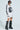 Dressed in the standout PUT A BOW ON IT JERSEY DRESS, a person showcases its sleek white oversized design, highlighted by a prominent number 5 and bold black details. With sunglasses, tattoos, and towering black boots embellished with chains, they boast an edgy attitude against the simple backdrop that enhances their daring look.