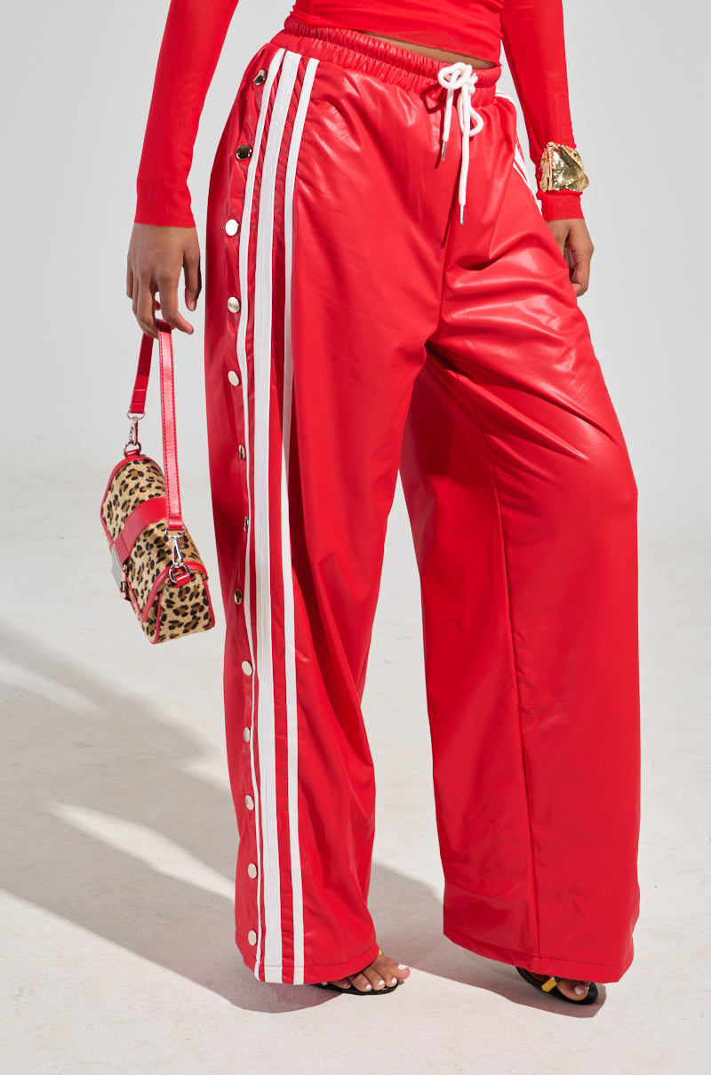 A person wearing the BELLERS PANT IN RED, featuring high-rise wide-leg snap button design with white stripes, pairs it with a matching red top and carries a leopard print handbag with red accents, channeling a 90s-inspired tearaway style.