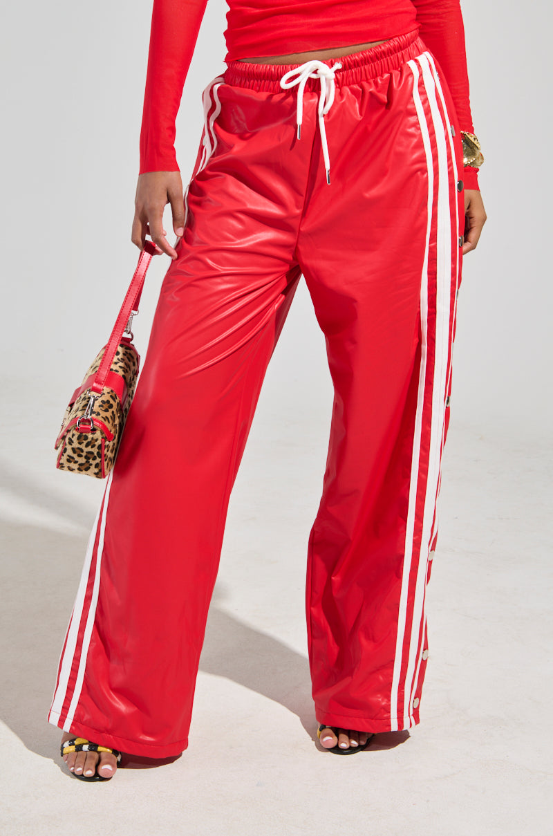 Wearing the BELLERS PANT IN RED, a person stands against a neutral background. The bright red track pants with white stripes evoke a 90's vibe, while they hold a leopard print bag in one hand, perfectly complementing their matching red top.