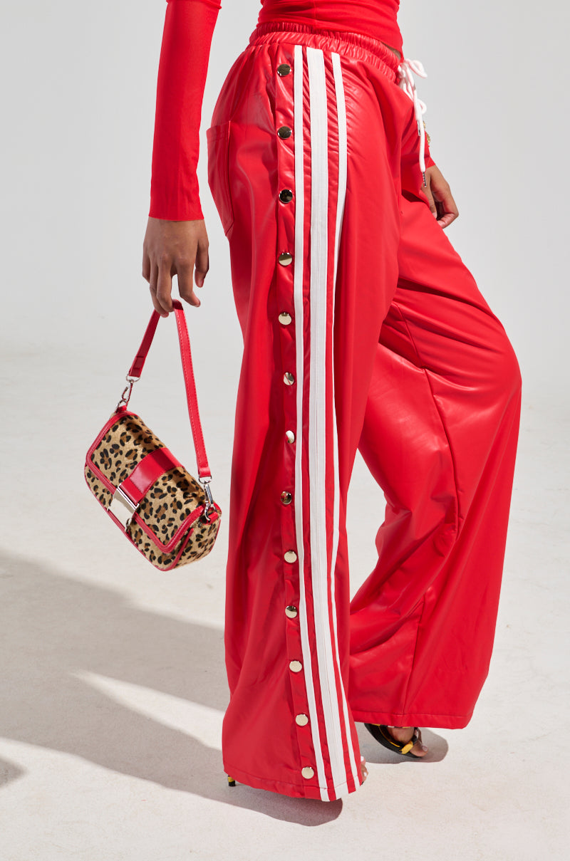 A person dressed in 90's-inspired BELLERS PANT IN RED, featuring tearaway snap-buttons and white stripes, with a coordinating red top, holds a leopard print handbag with red straps while standing on a light-colored surface.
