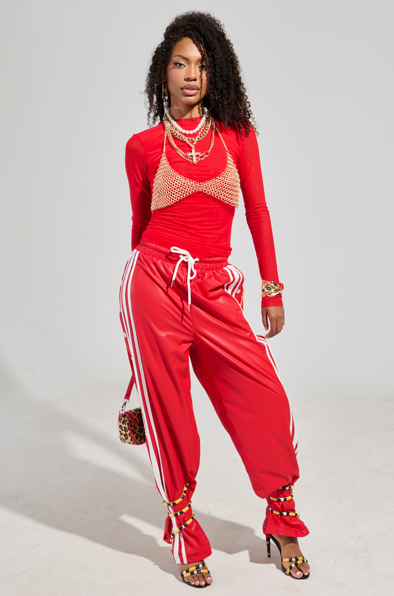 Dressed in a chic red outfit, the individual exudes confidence while wearing the BELLERS Pant in Red, which features white stripes reminiscent of 90’s-inspired tearaway styles. They complement it with a braided overlay top and strappy heels, accessorized with necklaces, bracelets, and a leopard print purse for added flair.