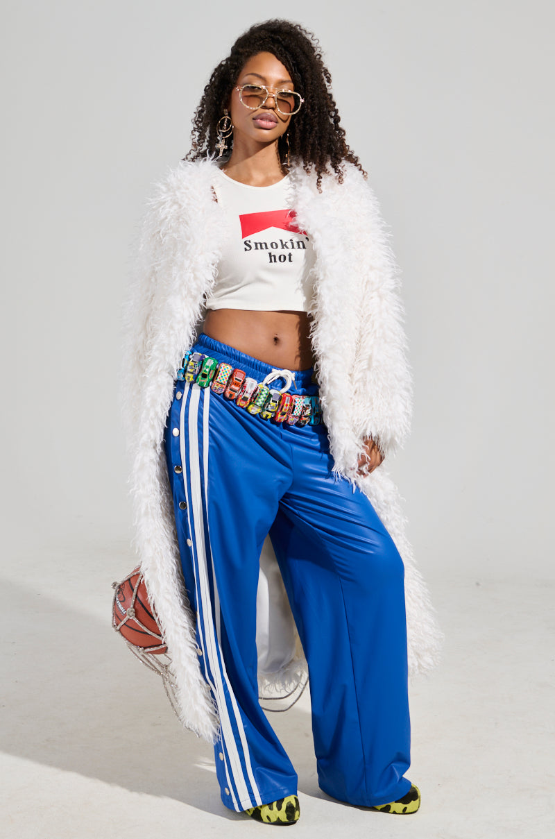 A woman poses confidently in a fluffy white coat and graphic crop top, while wearing the vibrant BELLERS PANT IN BLUE with white stripes. She accessorizes with sunglasses, hoop earrings, and a colorful belt. Channeling a 90’s-inspired vibe, a basketball is partially visible at her side.