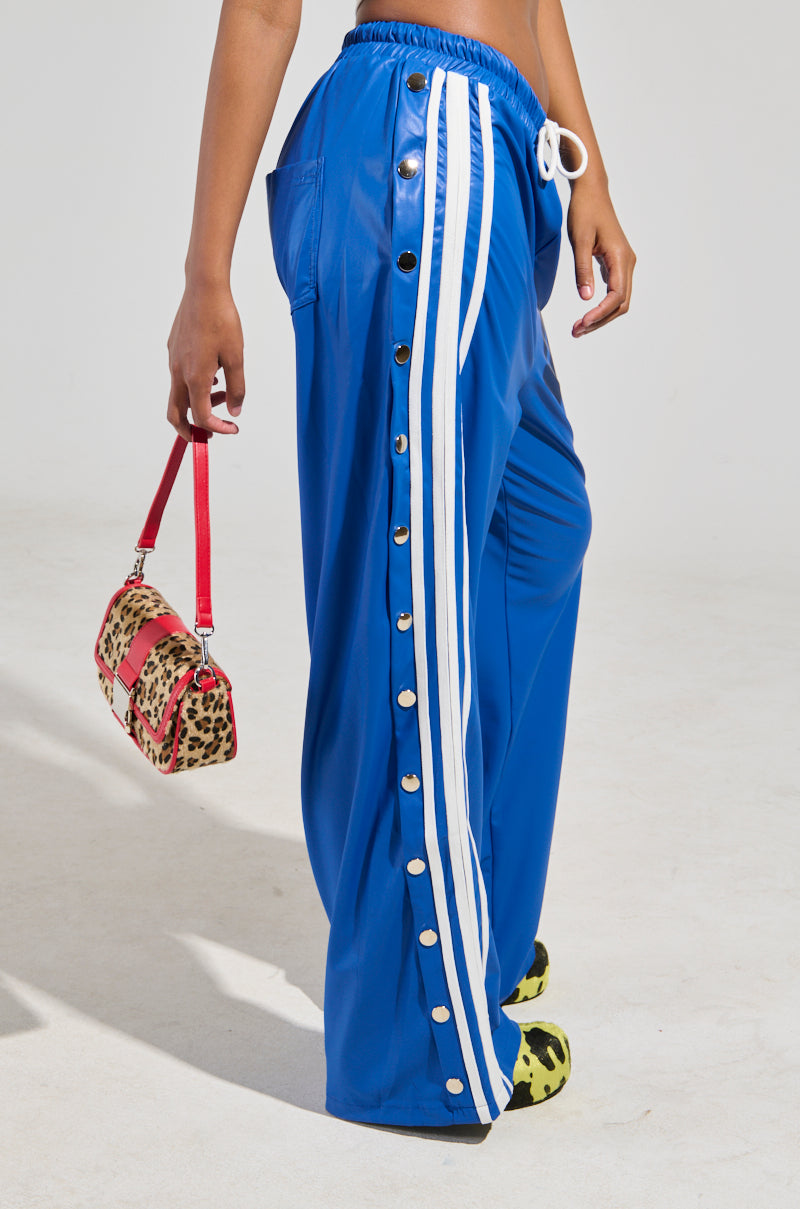A person dressed in the BELLERS PANT IN BLUE, 90’s-inspired snap-button track pants with white stripes and a drawstring, is holding a red leopard print handbag. They're also wearing patterned shoes. The background is plain white.