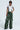 A person confidently stands against a plain background, wearing oversized green RUNNIN WILD CARGO PANT with a faux leather snakeskin pattern, paired with a white lace top featuring the number "07", large black sunglasses, and black shoes.