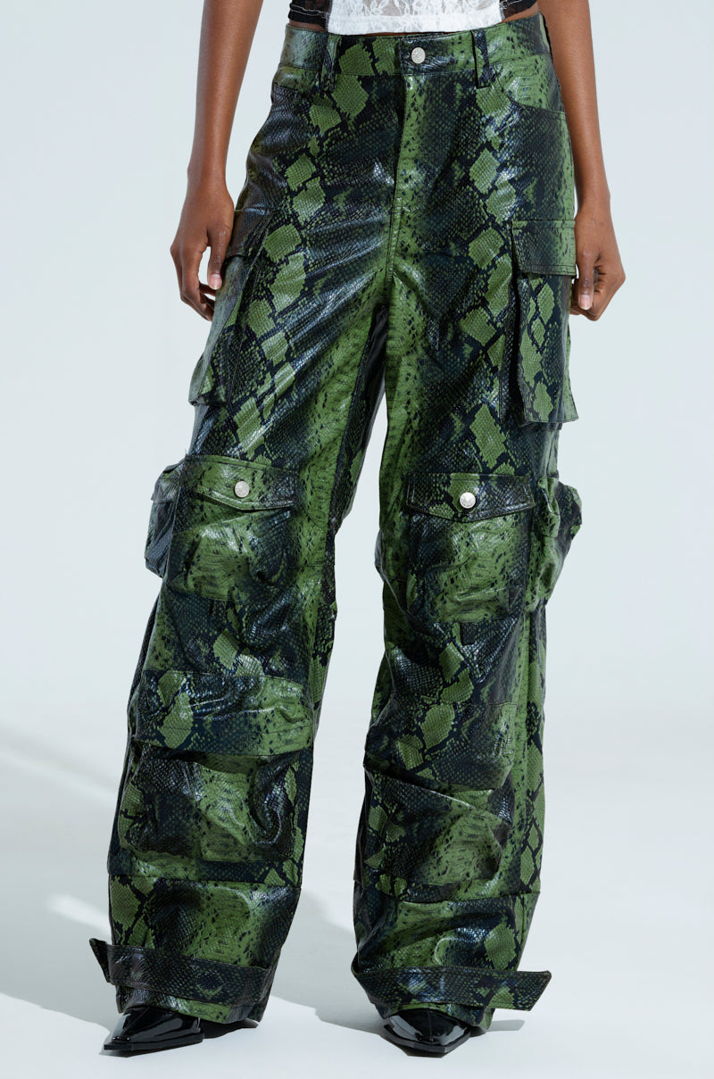 Person wearing the RUNNIN WILD CARGO PANT in a sleek green and black snakeskin pattern, featuring multiple pockets and button details. These faux leather pants boast a shiny, textured surface for a bold look.