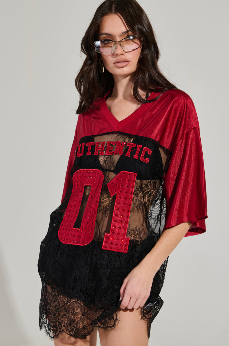 A person dressed in a red IT GIRL LACE JERSEY DRESS with "AUTHENTIC 01" printed on it, complete with a lace overlay. They have long, dark hair and are wearing sunglasses while posing against a neutral background—a perfect combination of elevated sportswear style.