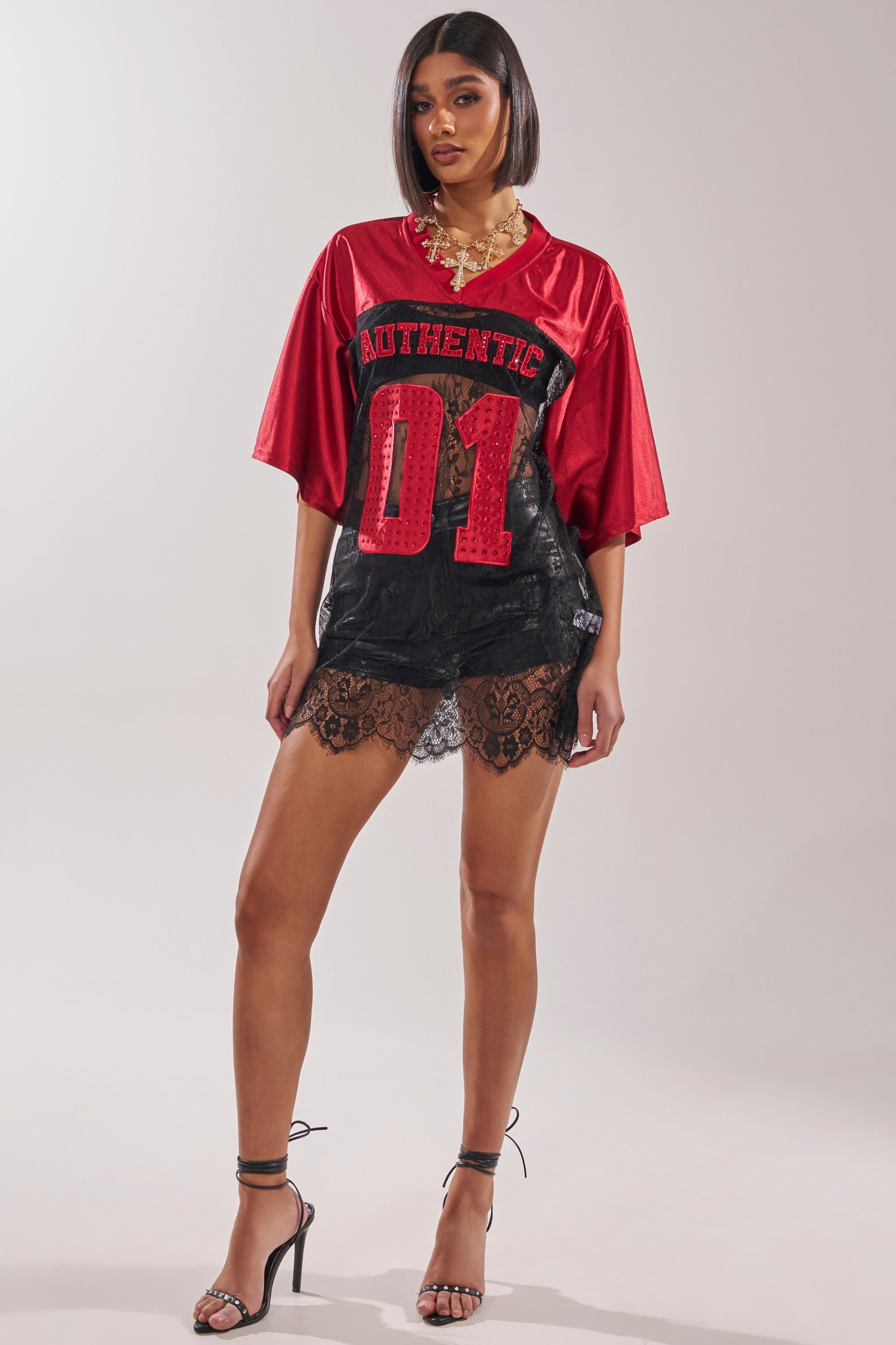 Woman models the IT GIRL LACE JERSEY DRESS—an oversized red jersey with "AUTHENTIC 01" text and black lace skirt—styled with black strappy heels, gold jewelry, and a sleek bob for an elevated sportswear look. 9