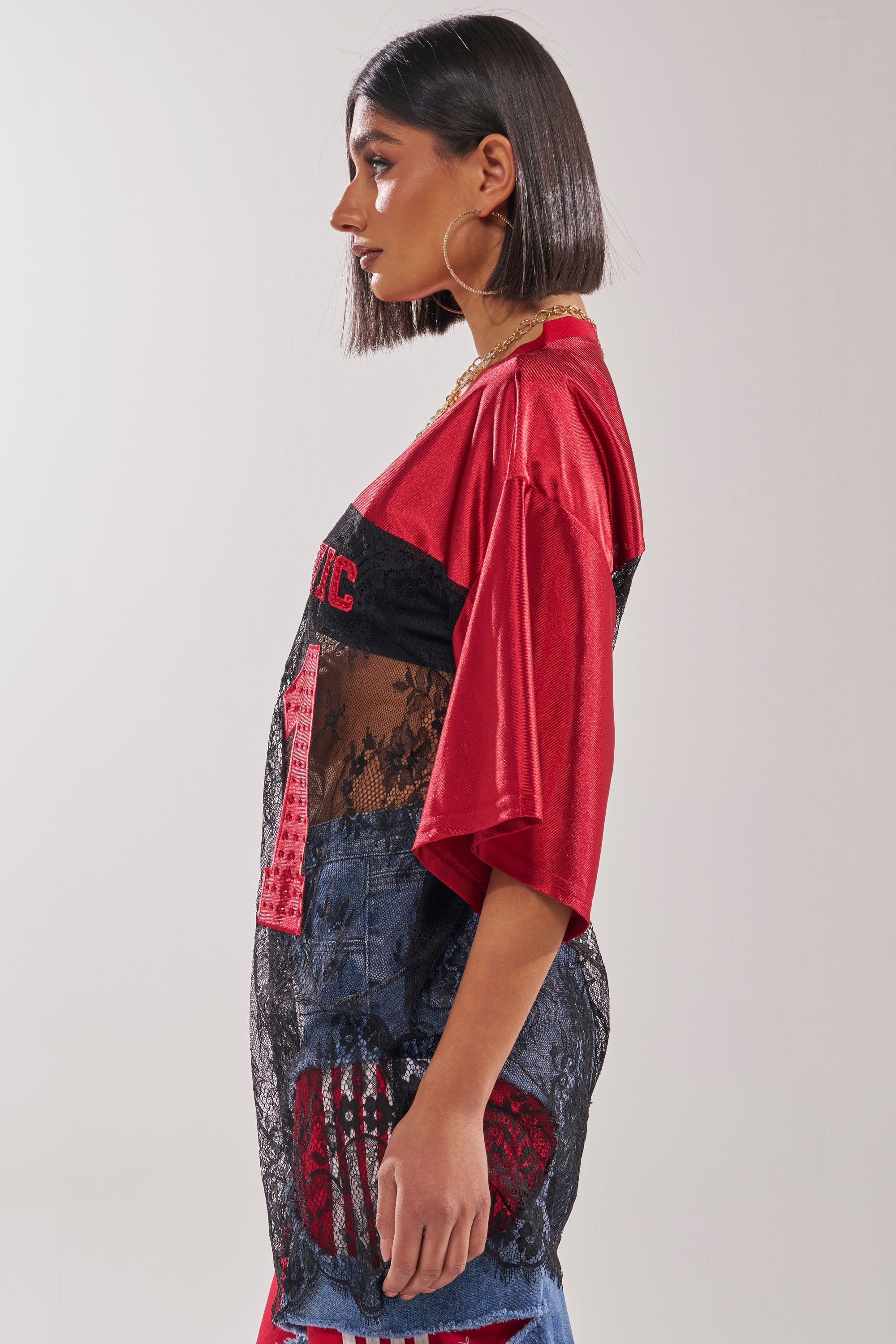 Wearing the IT GIRL LACE JERSEY DRESS, a woman with a sleek bob poses in profile. The dress features a red base with a sheer black lace overlay and blue denim details, styled with large hoop earrings against a plain light background. 10