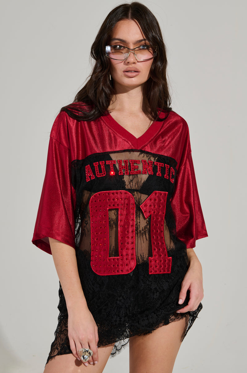 Dressed in an IT GIRL LACE JERSEY DRESS featuring a red top emblazoned with "Authentic 01" and a sheer black lace panel, a woman exudes confidence. Her long brown hair flows past her trendy sunglasses as she strikes a pose with one hand on her hip.