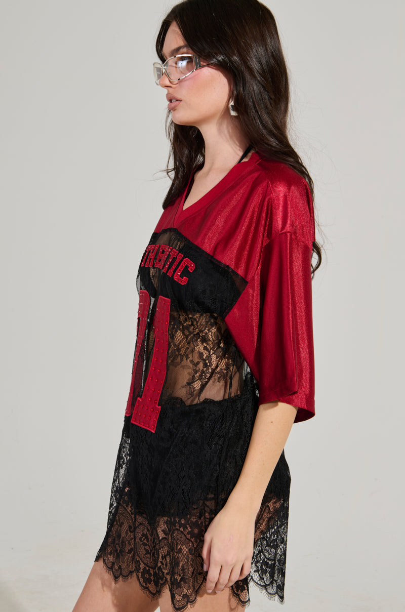 A woman with long dark hair wears glasses and the IT GIRL LACE JERSEY DRESS from AKIRA Label, featuring a red football jersey layered over a black lace dress for an elevated sportswear look. She stands against a plain background, gazing off to the side.