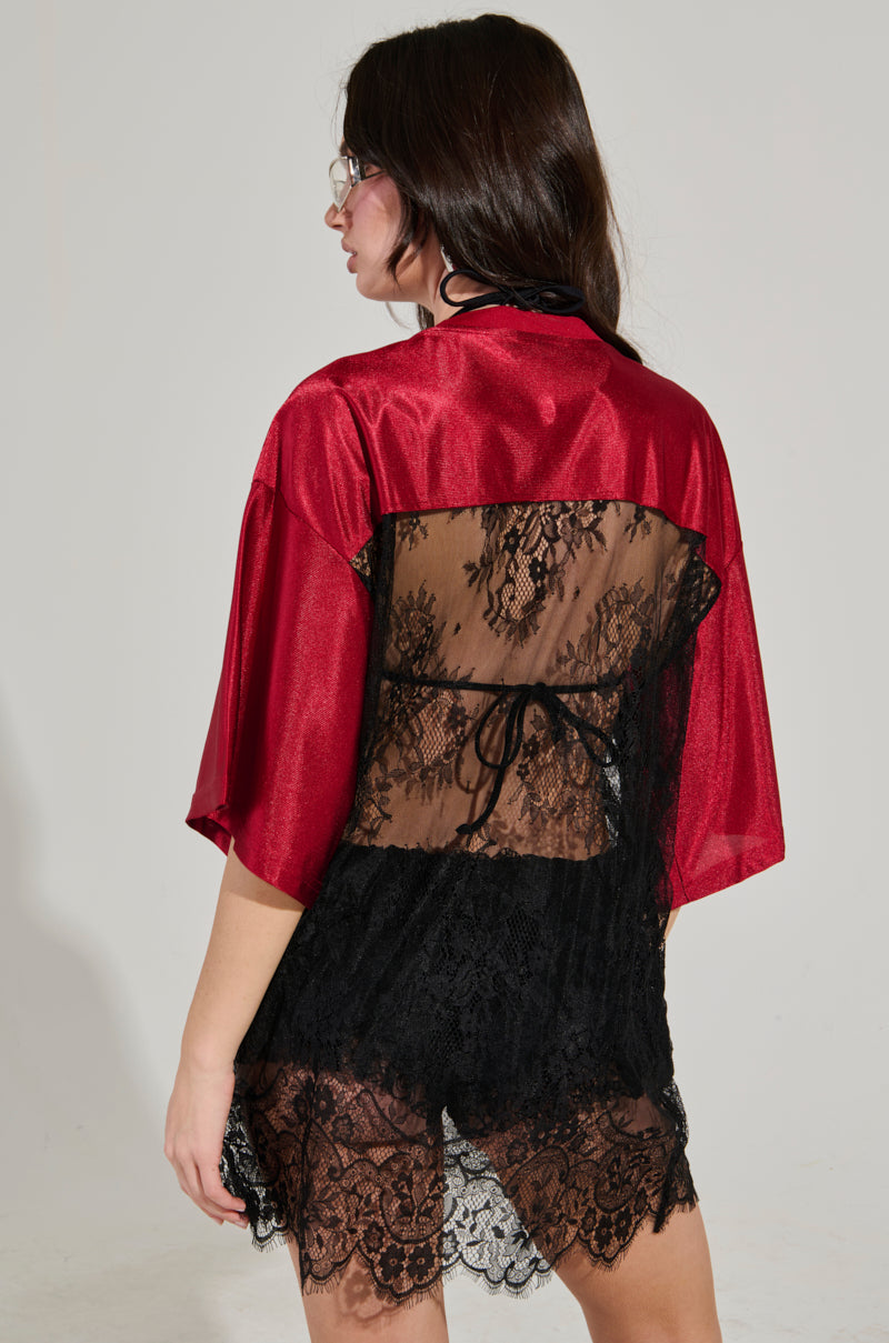 A woman with long dark hair wearing glasses is seen from the back, standing against a neutral background. She is wearing a red satin robe with black lace overlay reminiscent of the IT GIRL LACE JERSEY DRESS, featuring intricate floral patterns.