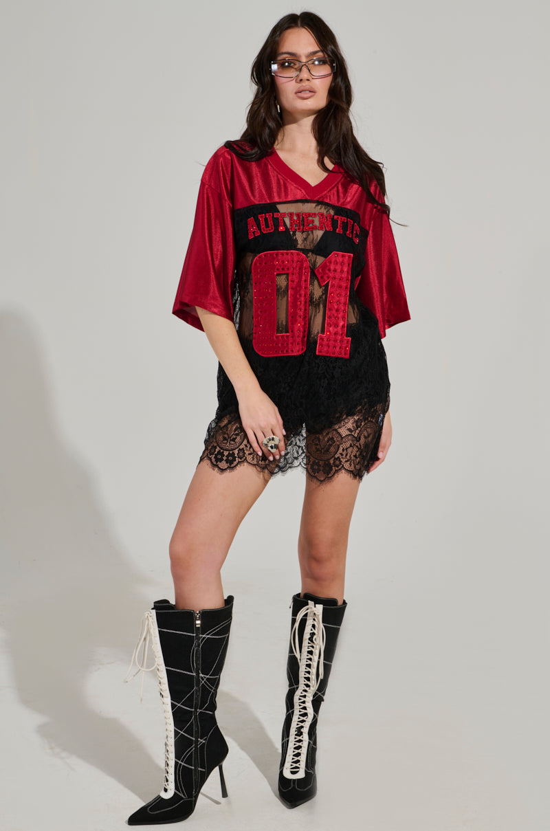 Wearing glasses, a confident individual stands out in an IT GIRL LACE JERSEY DRESS, a vibrant red piece with "AUTHENTIC 01" emblazoned on it. Paired with sleek black lace shorts and towering black boots adorned with striking white laces, they perfectly capture the essence of elevated sportswear against a simple background.
