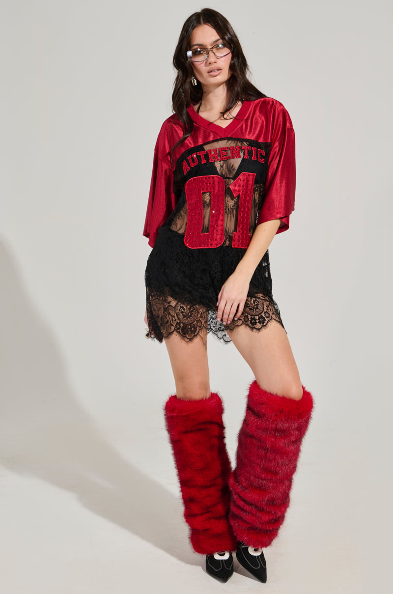 A woman confidently showcases elevated sportswear by wearing an IT GIRL LACE JERSEY DRESS, featuring a red jersey top with "Authentic 01" paired with a black lace skirt. Her look is completed with glasses, bold red fuzzy leg warmers, and black shoes, embodying the essence of an AKIRA Label Dress against a plain backdrop.