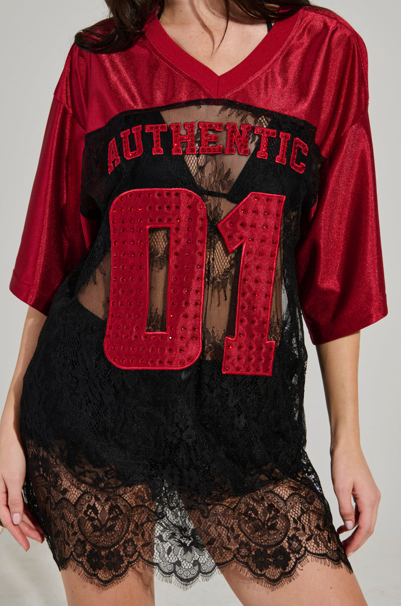 A person wearing an AKIRA Label IT GIRL LACE JERSEY DRESS in red and black, featuring "AUTHENTIC 01" and showcasing sheer lace detailing at the bottom, seamlessly blending elevated sportswear with elegance.