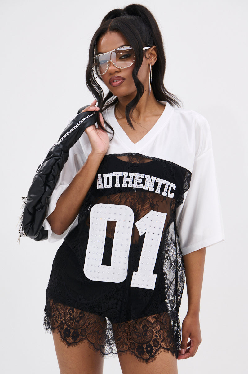 A woman in large sunglasses and hoop earrings poses confidently in the IT GIRL LACE JERSEY DRESS IN WHITE AND BLACK, a varsity-inspired mini dress with rhinestone-embellished lettering and sporty black lace detailing, holding a jacket over her shoulder.