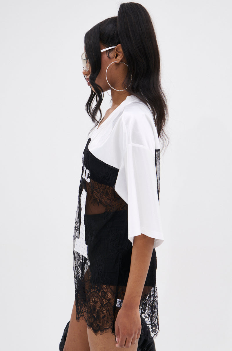 A woman with long dark hair in a high ponytail wears large hoop earrings, sunglasses, and the IT GIRL LACE JERSEY DRESS IN WHITE AND BLACK over a white oversized tee, serving varsity-inspired mini dress vibes against a plain background.