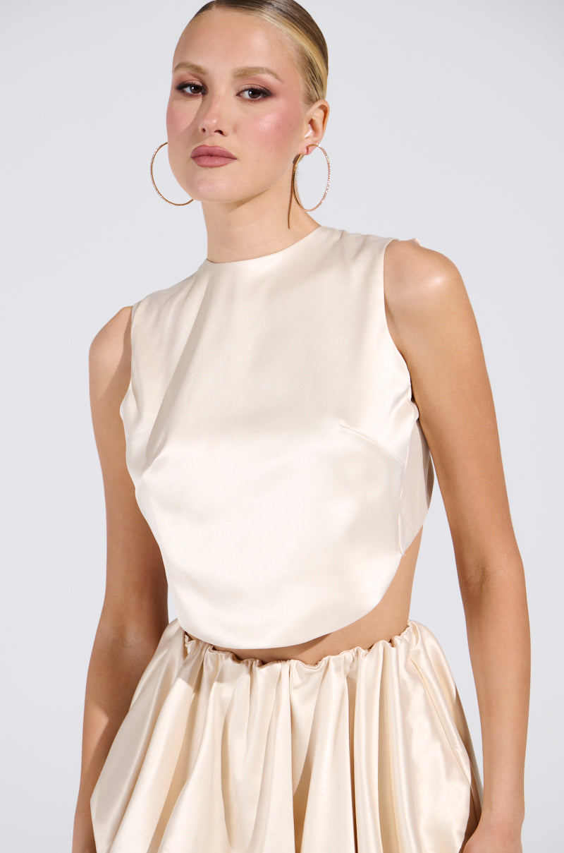A woman wears the CITY POPPIN CROPPED OPEN BACK BLOUSE in cream satin, styled with hoop earrings. Her slicked-back hair enhances her elegant pose against a plain, light background.