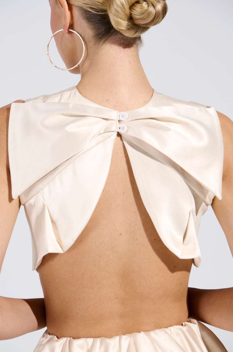 Facing away from the camera, an individual shows off an elegant cream CITY POPPIN CROPPED OPEN BACK BLOUSE with a large bow detail. Their chic look is enhanced by blonde hair in a bun and large hoop earrings, radiating effortless grace.