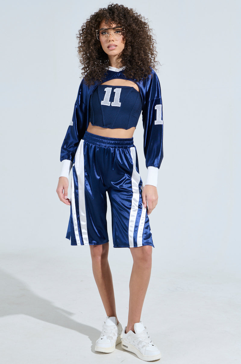 A person with curly hair confidently wears a blue crop top numbered 11 and pairs it with sporty-chic AKIRA Label GAME TIME SHORTS. They complete the look with white sneakers and clear-framed glasses, standing against a plain light background.