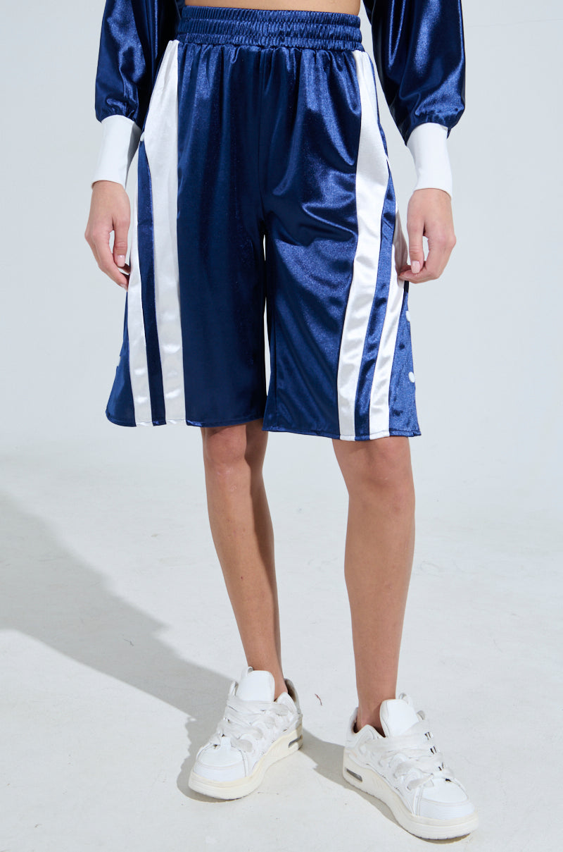 Wearing GAME TIME SHORTS by AKIRA Label in blue satin with white side stripes, paired with a matching long-sleeve top and white sneakers, this person gives off a sporty-chic vibe on a light-colored floor.