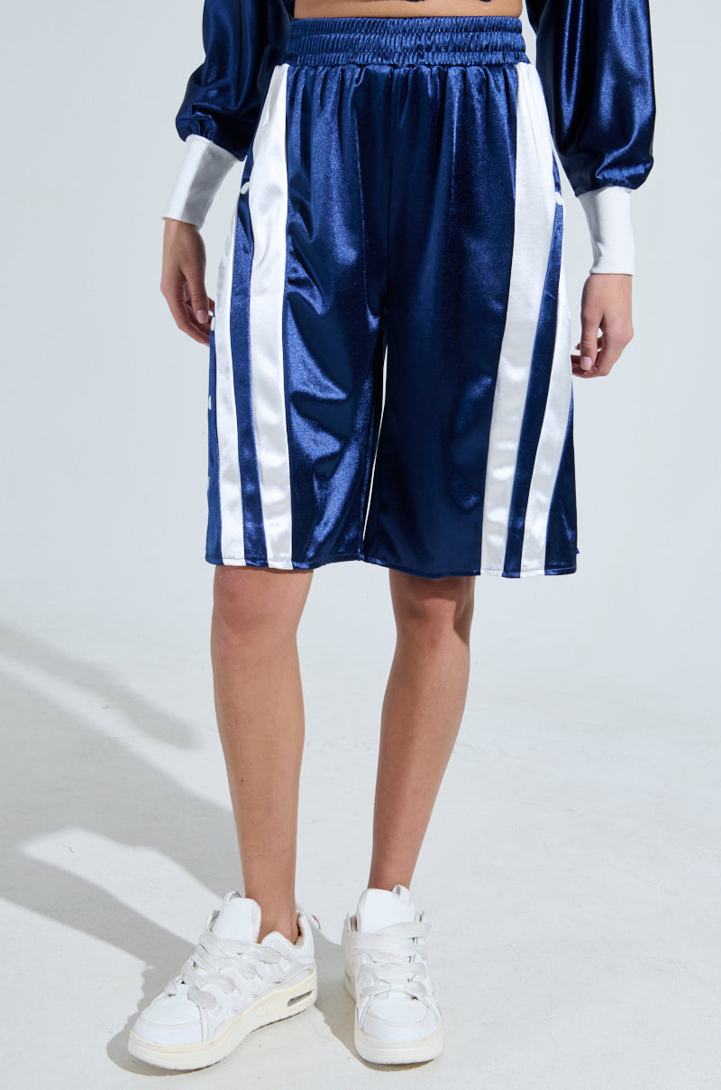 A person stands in a studio wearing AKIRA Label's GAME TIME SHORTS, featuring a sporty-chic design in shiny blue with white stripes. They're paired with a matching long-sleeve top and crisp white sneakers against a plain white backdrop.