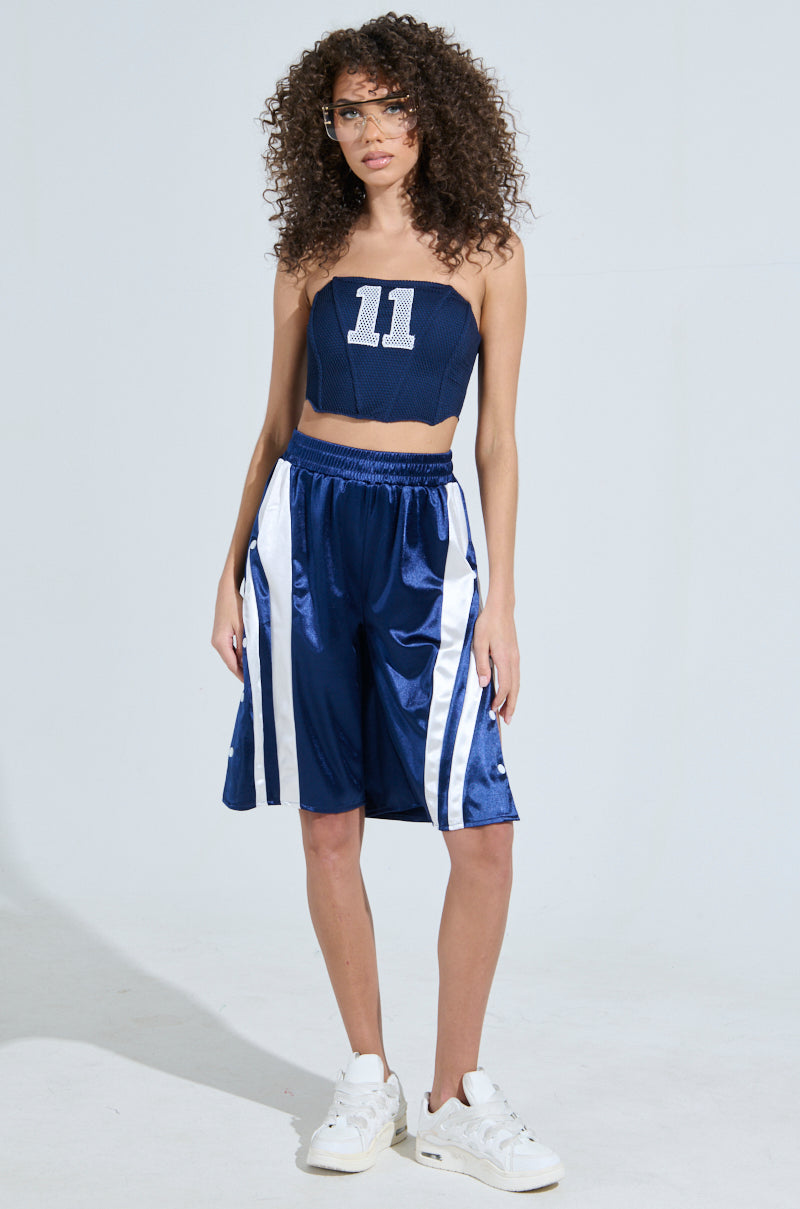 A person with curly hair confidently wears a dark blue AKIRA Label sports outfit, featuring GAME TIME SHORTS adorned with white stripes and a sleeveless top displaying the number 11. Their sporty-chic style is completed with white sneakers against a light background.