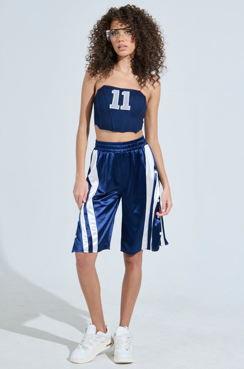 A curly-haired woman poses in a sporty-chic look, with a sleeveless crop top featuring the number 11 and GAME TIME SHORTS by AKIRA. She finishes her outfit with white sneakers and sunglasses against a plain white backdrop.