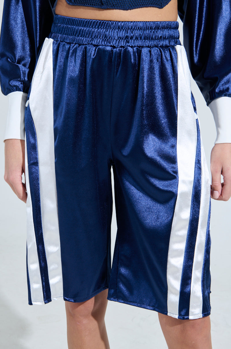 A person in sporty-chic bottoms with shiny navy fabric and wide white side stripes. The GAME TIME SHORTS from AKIRA feature an elastic waistband, sitting above the knees, on a plain light gray background.