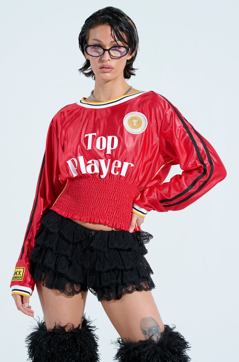 A person in a red sporty TOP PLAYER SCRUNCH JERSEY, black ruffled shorts with a ruched waistband, fuzzy leg warmers, and sunglasses poses confidently against a light background, showcasing their short edgy hairstyle.