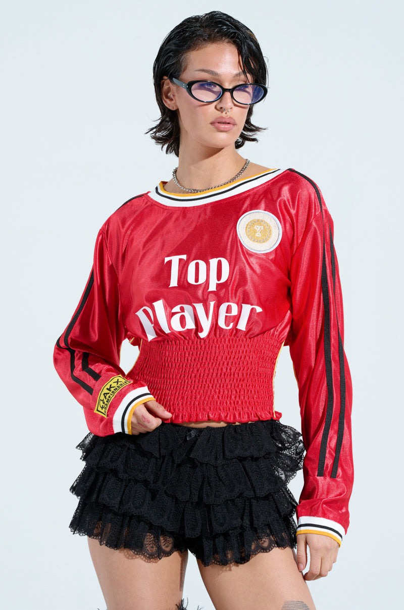Someone with short dark hair wears sporty glasses, a red TOP PLAYER SCRUNCH JERSEY with gold patches and black stripes, and a black lace ruffled skirt with a ruched waistband, all set against a light blue background.