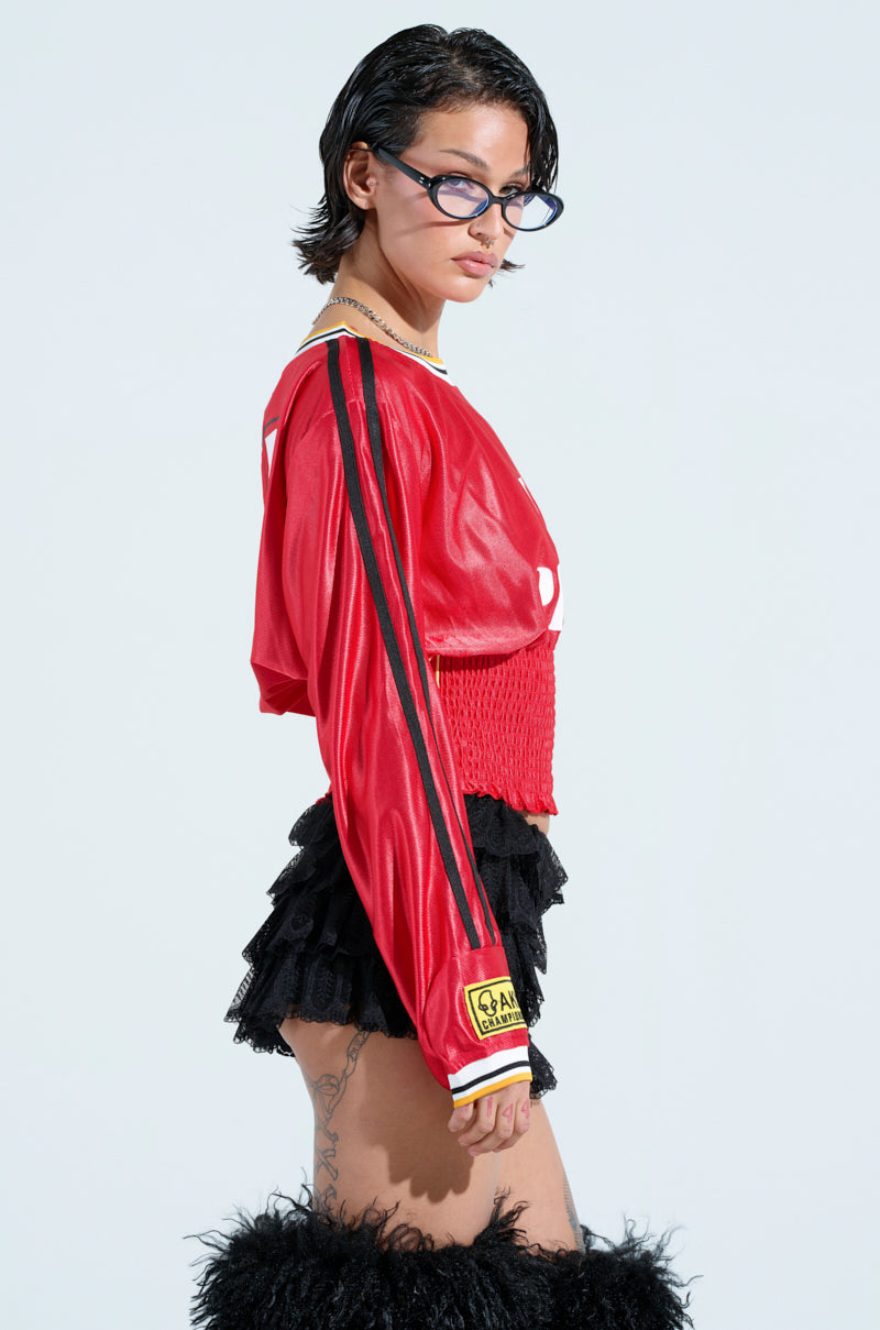Sporting the Top Player Scrunch Jersey in red with black stripes, they pair it with a layered black skirt for a waist-cinching silhouette and finish with black furry knee-high boots. With short dark hair, sunglasses, and leg tattoos against a light backdrop, they exude effortless cool.