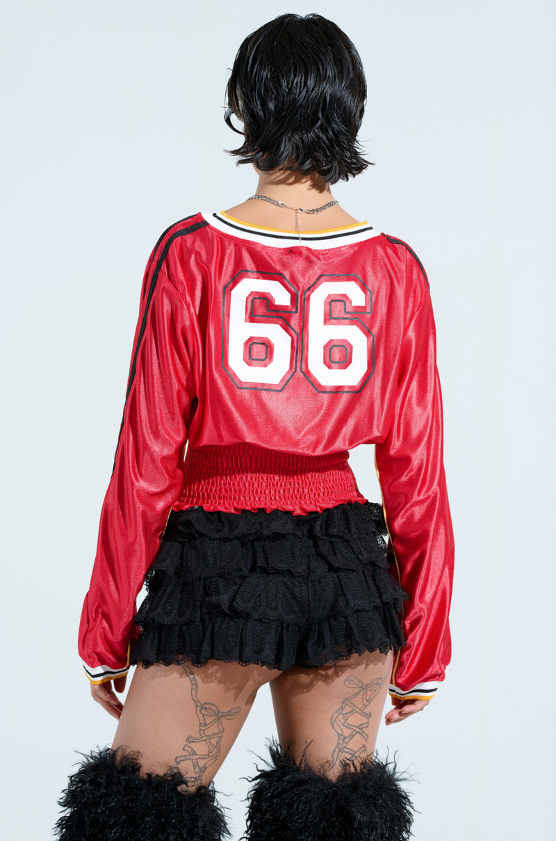 A person with short hair is wearing a red TOP PLAYER SCRUNCH JERSEY, number 66 on the back, paired with a black ruffled skirt with a ruched waistband and black leg warmers. They have tattoos on their legs and stand against a plain background.