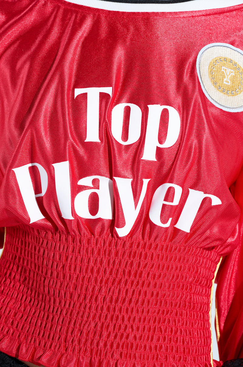 The TOP PLAYER SCRUNCH JERSEY combines sporty-chic style with a waist-cinching silhouette, featuring "Top Player" in bold white letters across the chest. A round patch with a yellow border and emblem is stitched near the top right, complementing the ruched waistband for added flair.