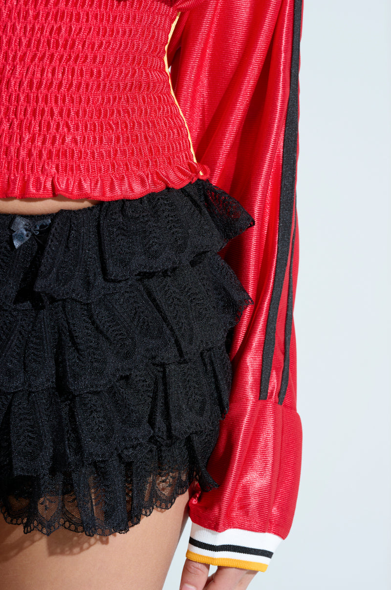 Wearing the red TOP PLAYER SCRUNCH JERSEY with black vertical-striped long sleeves and textured detailing, paired with frilly black lace shorts, creates a waist-cinching look with a slight midriff on a light background.
