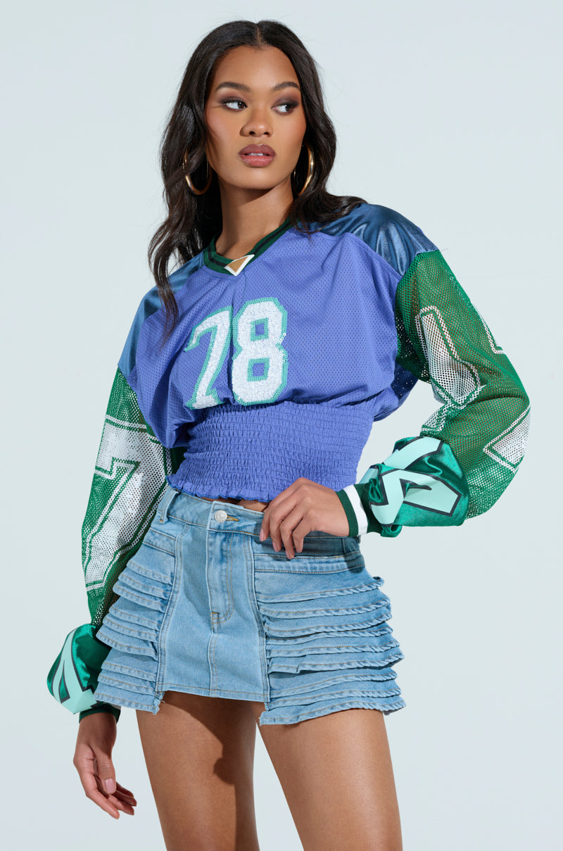 A woman in a blue and green sports jersey labeled 78, paired with a tiered denim skirt, stands against a light background. Her long hair and hoop earrings complete the look reminiscent of the MEXICO RUCHED JERSEY style.