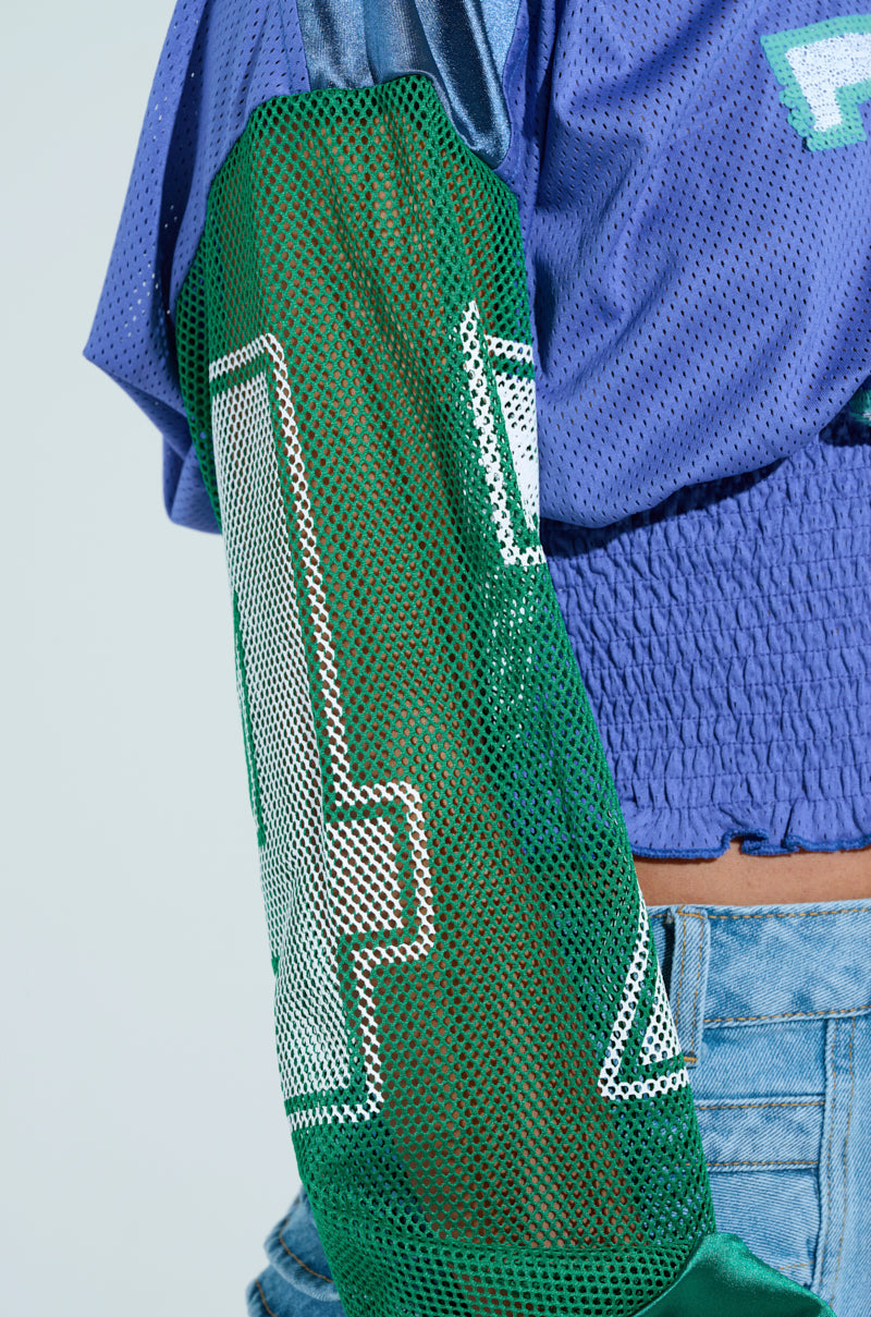 A close-up of a person wearing the MEXICO RUCHED JERSEY with a color-block design, cropped blue section, and green mesh sleeve featuring the white number 17. They effortlessly pair it with high-waisted light blue jeans.