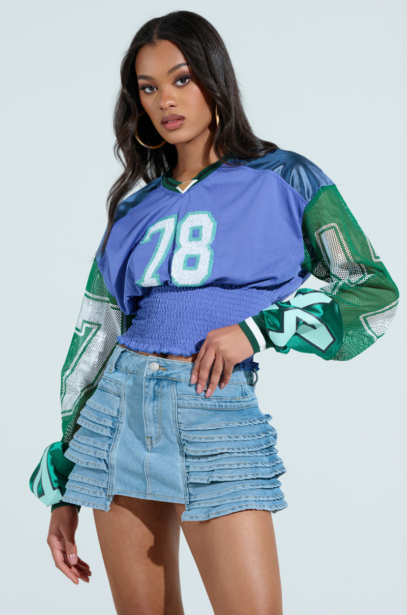 A woman confidently poses against a light background in a color-blocked MEXICO RUCHED JERSEY, featuring blue and green with the number 78. She pairs it with a layered ruffle denim skirt, completing her chic look with long dark hair and hoop earrings.