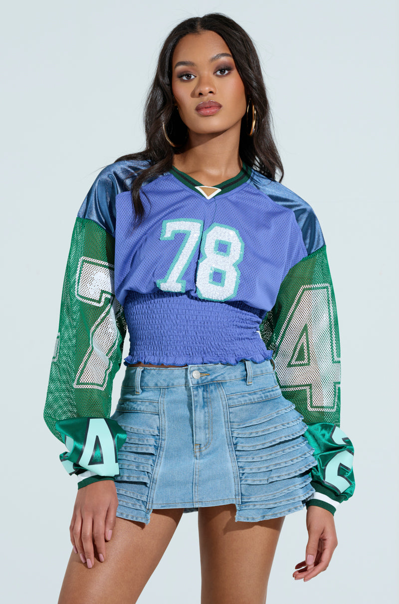 A woman confidently poses against a plain backdrop in the blue and green MEXICO RUCHED JERSEY featuring number 78, paired with a ruffled denim mini skirt. Her long hair falls down, complemented by hoop earrings.